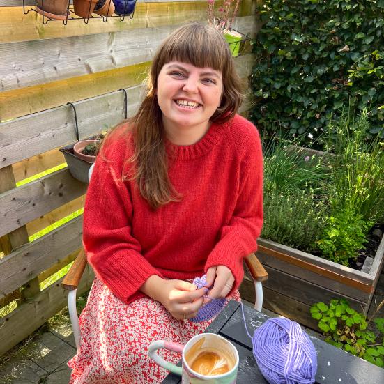Maria sidder ved et havebord, mens hun strikker fra et lilla garnnøgle. På bordet står et keramikkrus med kaffe.