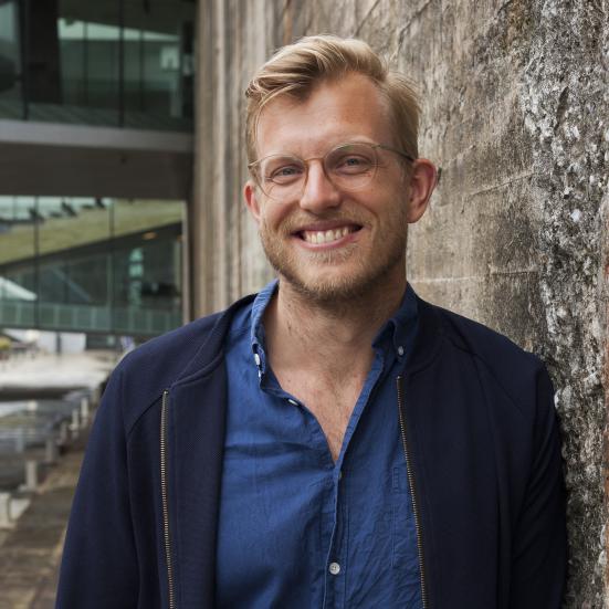 Nils Valdersdorf Jensen kigger ind i kameraet med et stort smil. Han står foran en grålig murstensvæg.