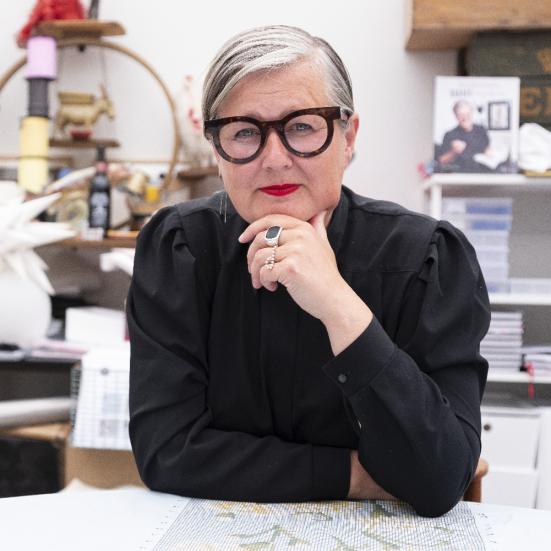 Trine ser direkte ind i kameraet med en hånd mod hagen. Hun er iført en sort bluse og markante briller.