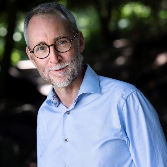 Tom Buk-Swienty kigger ind i kameraet med et let smil på læberne. Han er iført en blå skjorte. I baggrunden aner man en allé med høje træer.