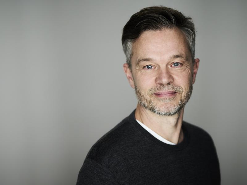 Portrætfoto af Jesper Bugge Kold i farver. Han kigger ind i kameraet med et lille smil på læberne og er iført en sort bluse.