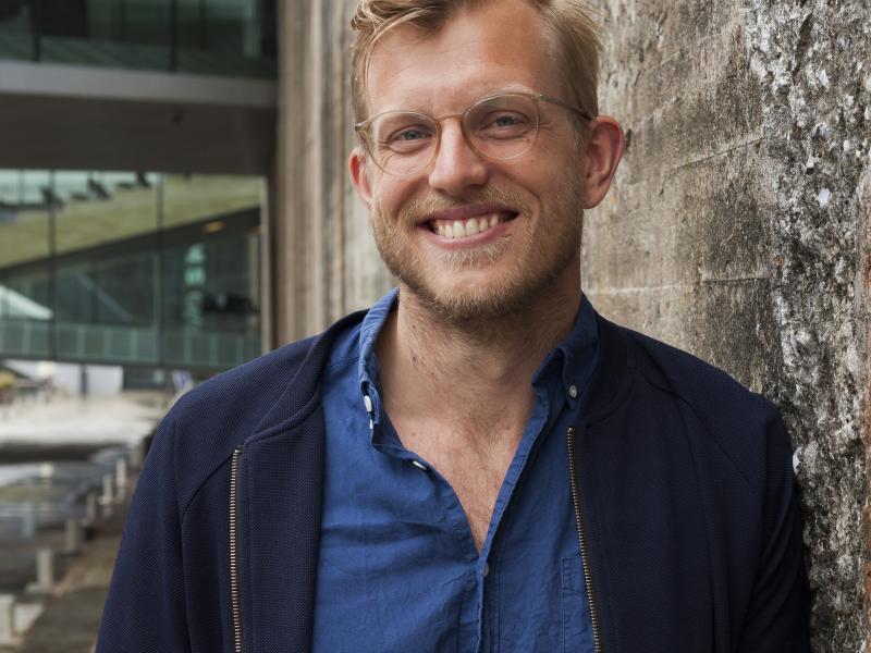 Nils Valdersdorf Jensen kigger ind i kameraet med et stort smil. Han står foran en grålig murstensvæg.
