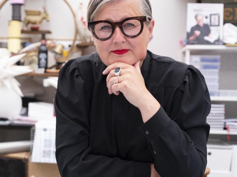 Trine ser direkte ind i kameraet med en hånd mod hagen. Hun er iført en sort bluse og markante briller.