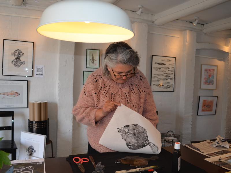 Linea/Momlis står ved et bord og løfter et fisketryk op, så man kan se motivet.