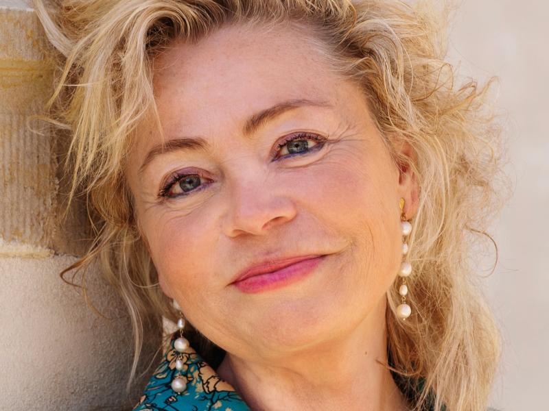 Annette Bjergfeldt står lænet mod en beige mur. Hun er iført en blåmønstret jakke og perleøreringe og smiler mod kameraet.