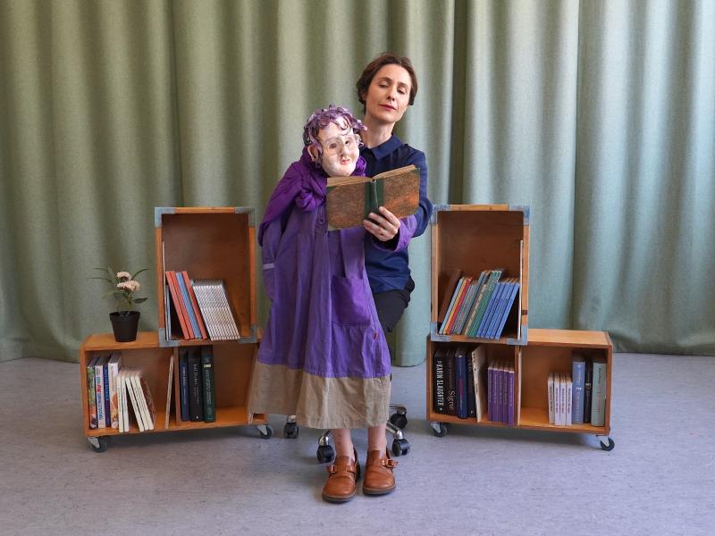 Foto af skuespiller og dukke på scenen mellem bøger