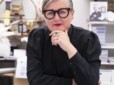 Trine ser direkte ind i kameraet med en hånd mod hagen. Hun er iført en sort bluse og markante briller.