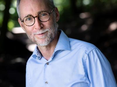 Tom Buk-Swienty kigger ind i kameraet med et let smil på læberne. Han er iført en blå skjorte. I baggrunden aner man en allé med høje træer.