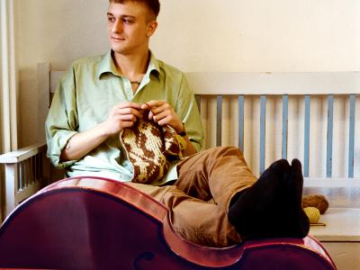 Albert sidder på en slagbænk og kigger ud ad vinduet, mens han strikker. Foran ham ligger en kontrabas, som han hviler fødderne på.