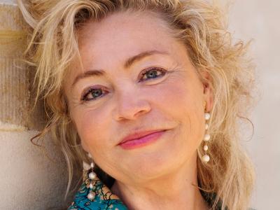 Annette Bjergfeldt står lænet mod en beige mur. Hun er iført en blåmønstret jakke og perleøreringe og smiler mod kameraet.