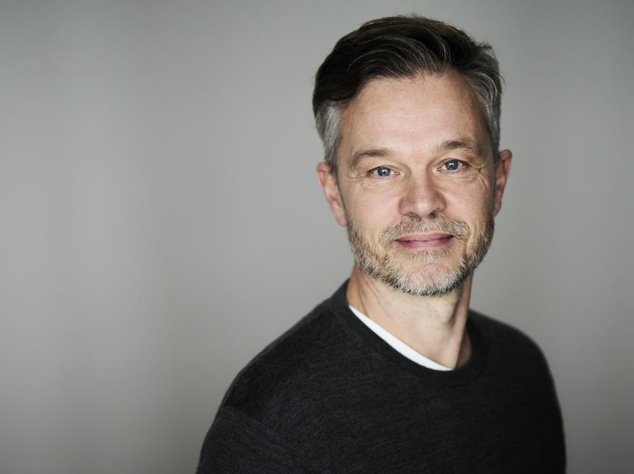 Portrætfoto af Jesper Bugge Kold i farver. Han kigger ind i kameraet med et lille smil på læberne og er iført en sort bluse.