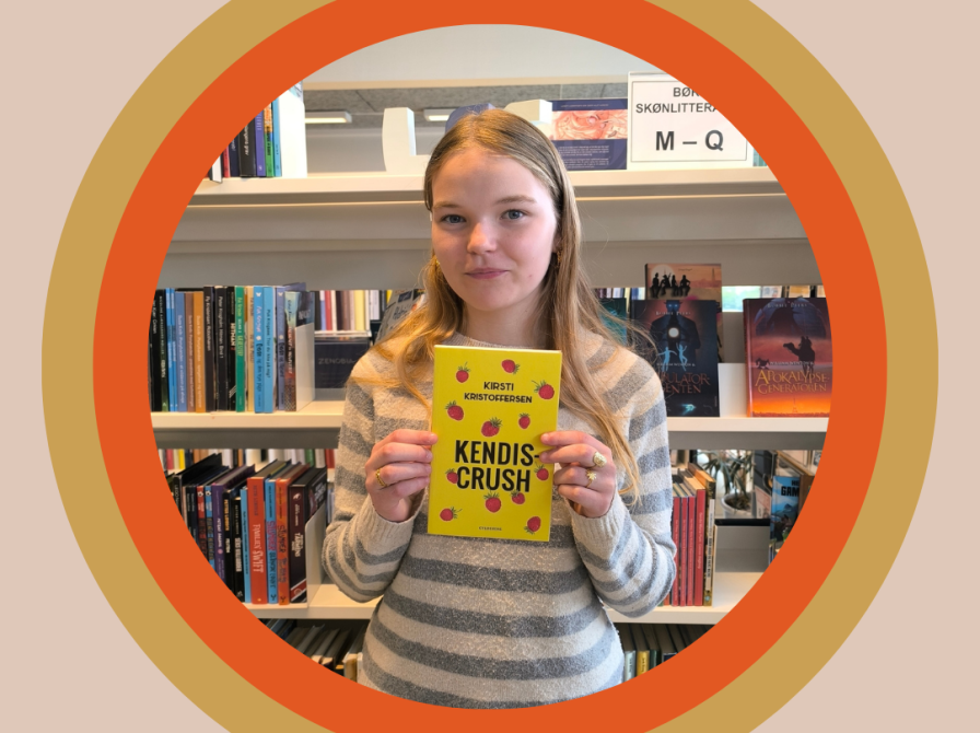 Clara har bogen "Kendiscrush" i hænderne. Hun står foran en bogreol.